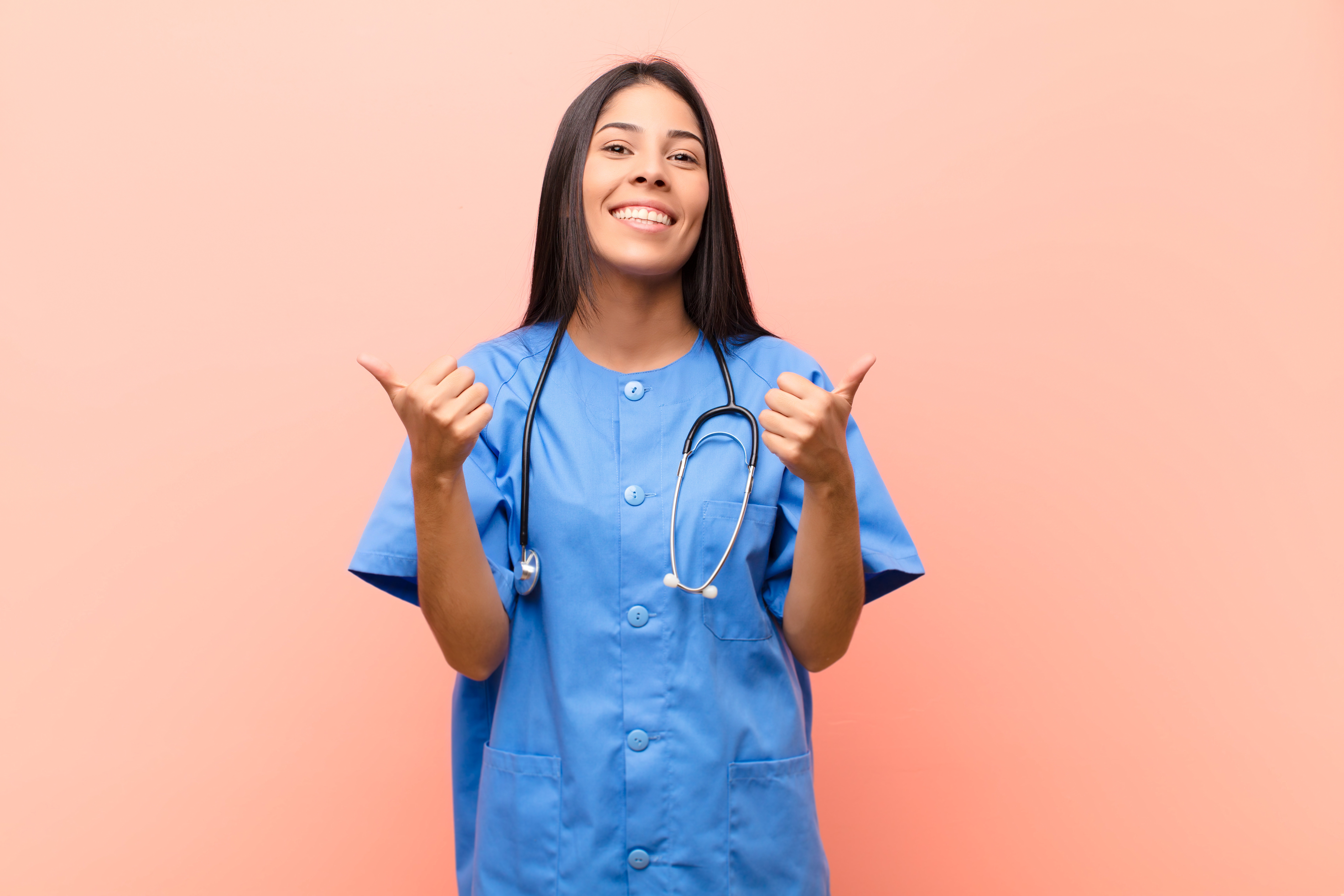 young latin nurse smiling joyfully and looking happy, feeling carefree and positive with both thumbs up against pink wall