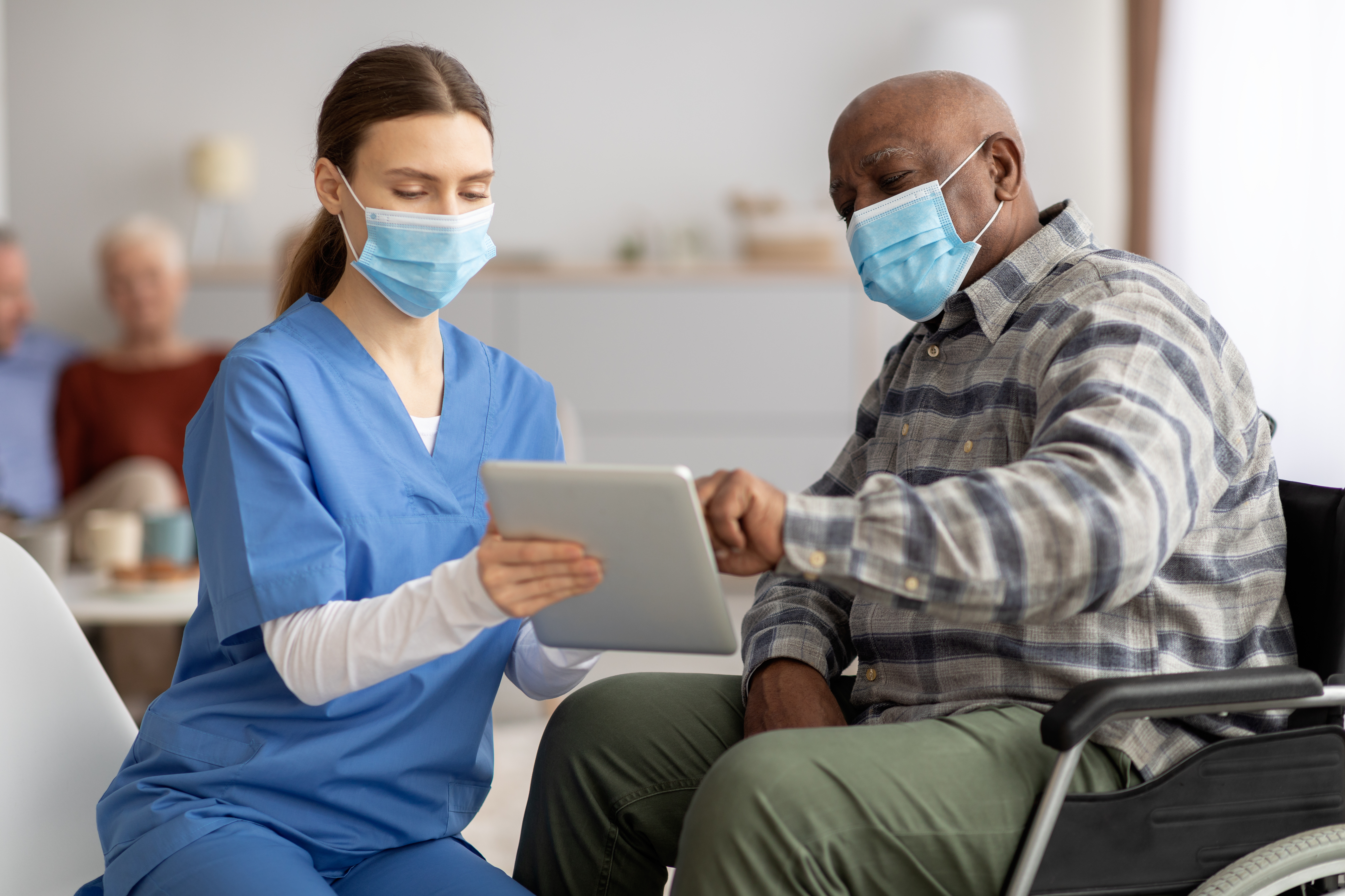 Woman in workwear nurse helping black senior man in weheelchair using digital tablet, elderly african american male patient surfing on Internet at nursing home, wearing protective face masks